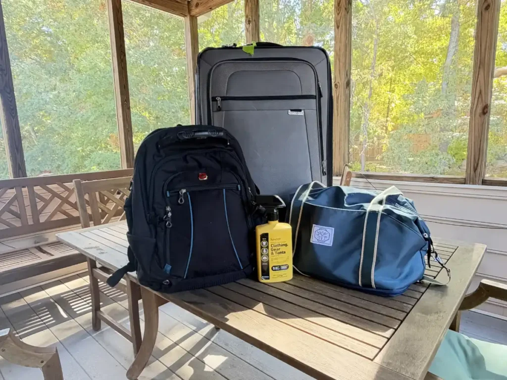 Gray upright rolling suitcase, black Wenger SwissGear backpack, and blue duffel bag arranged on an outdoor wooden table alongside a yellow spray bottle of Sawyer Permethrin insect repellent.